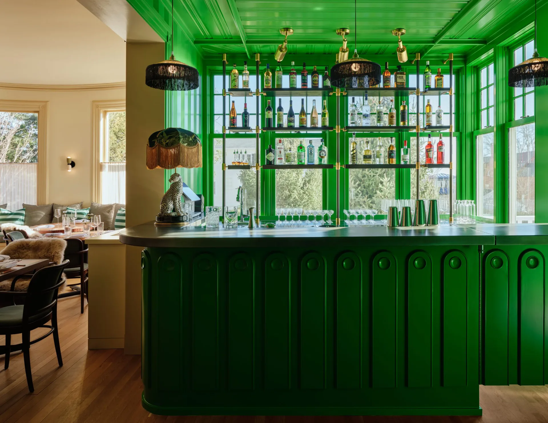A striking green bar interior with floor-to-ceiling cabinetry, glass shelving, pendant lighting, and warm wood flooring, designed by Jun Chun.
