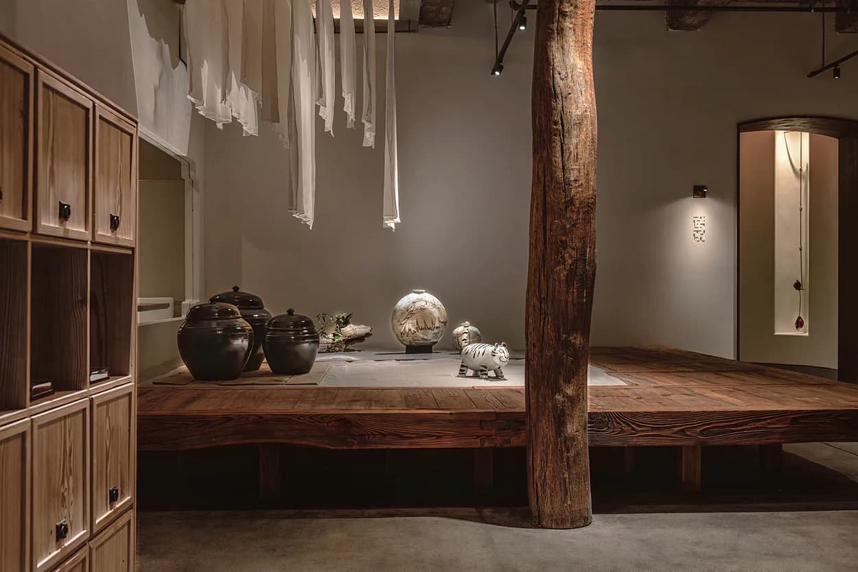 Contemporary gallery interior with wooden display platform, rustic timber column, neutral palette, and minimalist ceramic vessels by Junho Choi.