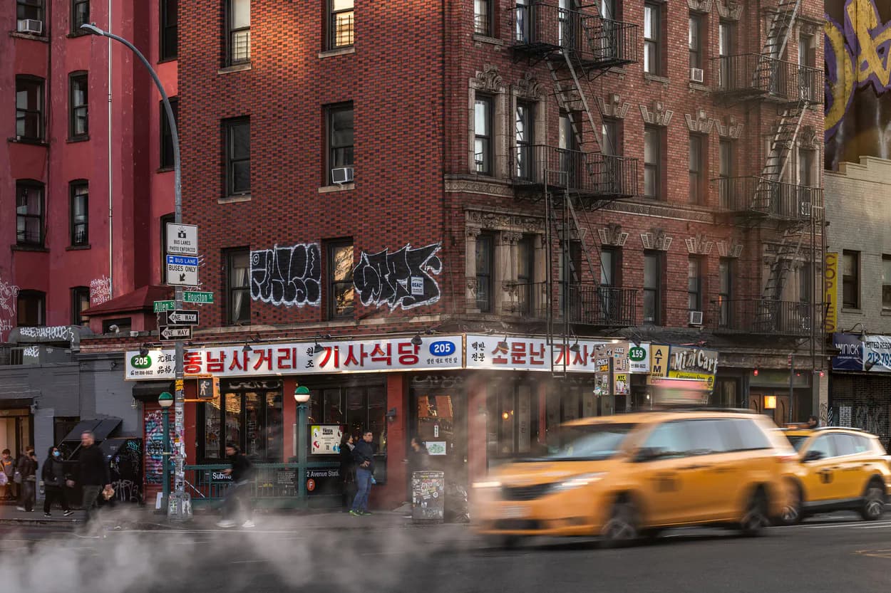 Urban street scene featuring red brick buildings with storefront signage and a yellow taxi in motion, architectural photography by Junho Choi.