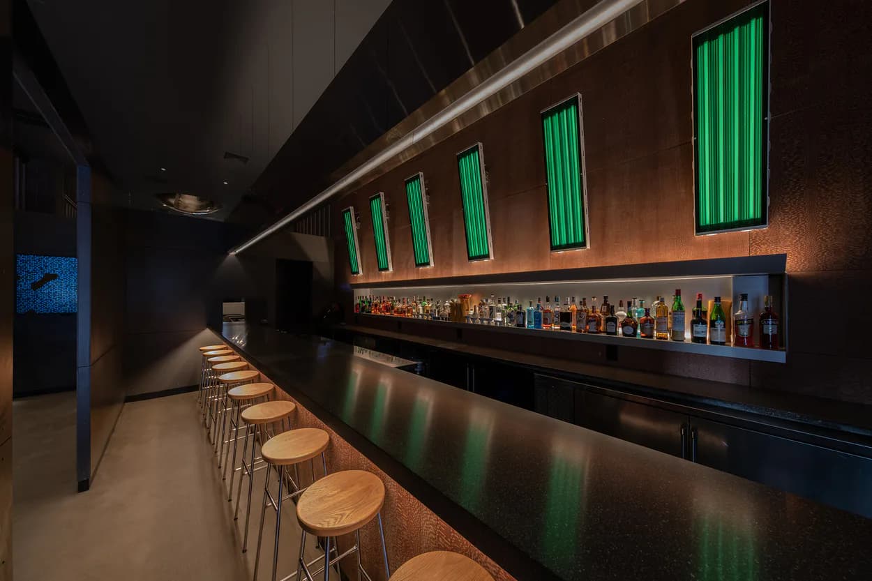 Contemporary bar interior with wooden counter stools, dark cabinetry, illuminated green neon vertical panels, and ambient lighting by Junho Choi.