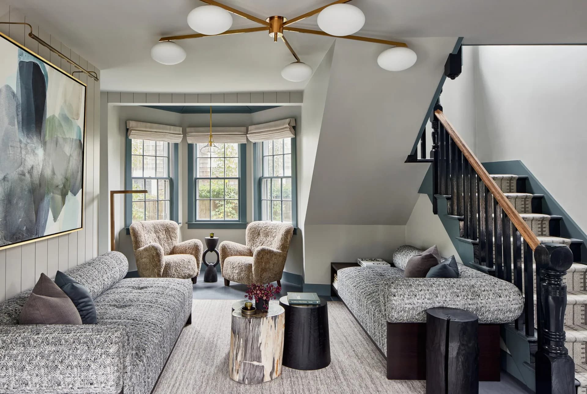 Modern residential interior with two gray upholstered seating areas flanking a bay window, brass ceiling fixture, and black staircase by Kaitlin McQuaide.