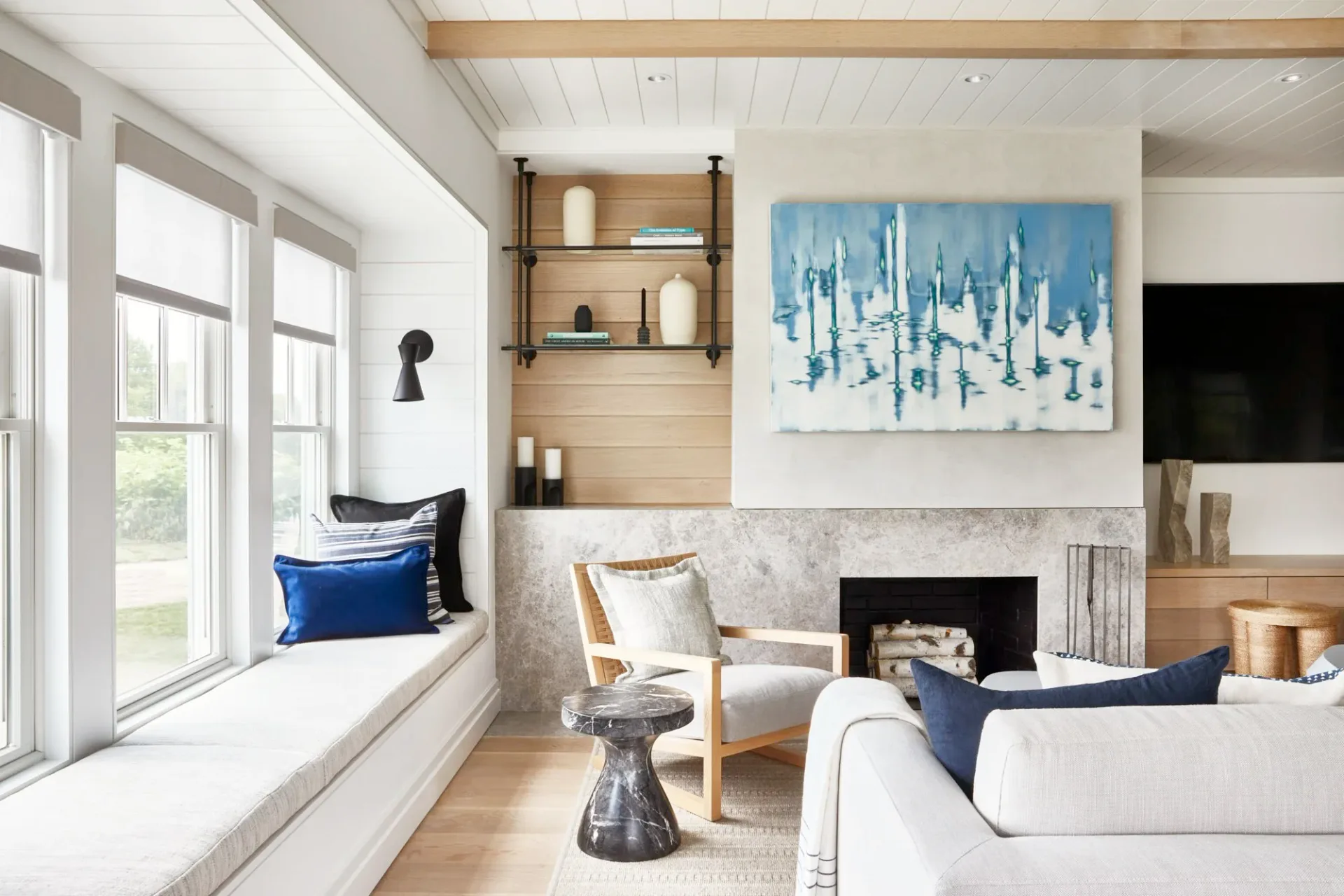 Bright living room with white shiplap fireplace, blue abstract artwork, window seat with navy pillows, light wood shelving, and modern furnishings by Kaitlin McQuaide.