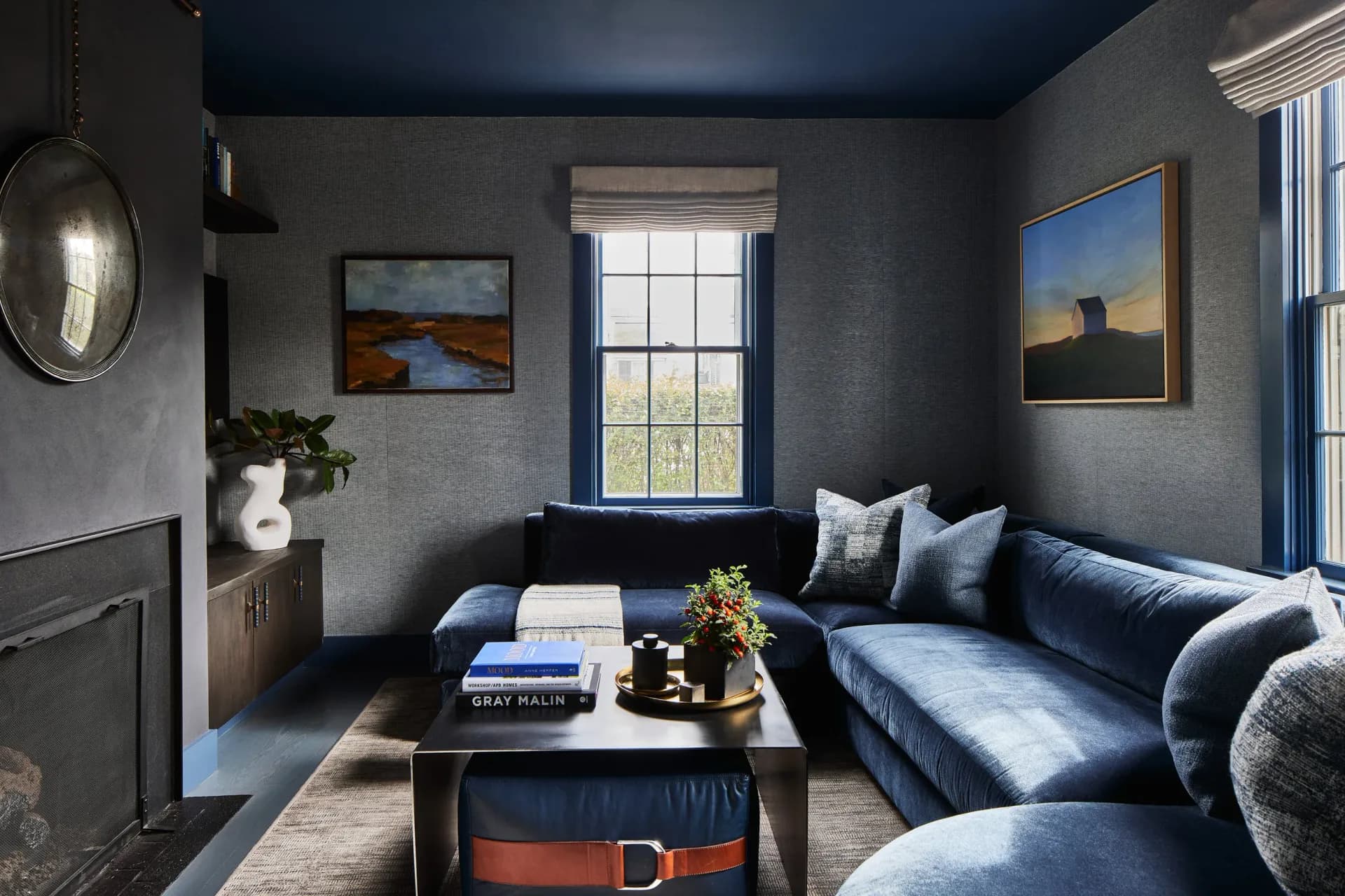 A contemporary living room with blue upholstered sectional sofa, dark gray textured walls, fireplace, and large windows overlooking landscape, designed by Kaitlin McQuaide.