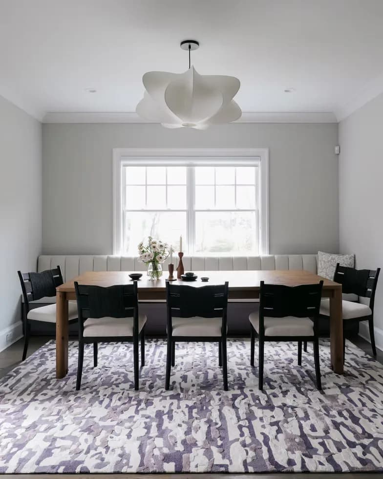 Modern dining room with natural wood table, black chairs, gray patterned rug, and sculptural pendant light overlooking large windows by Kanika Gupta.