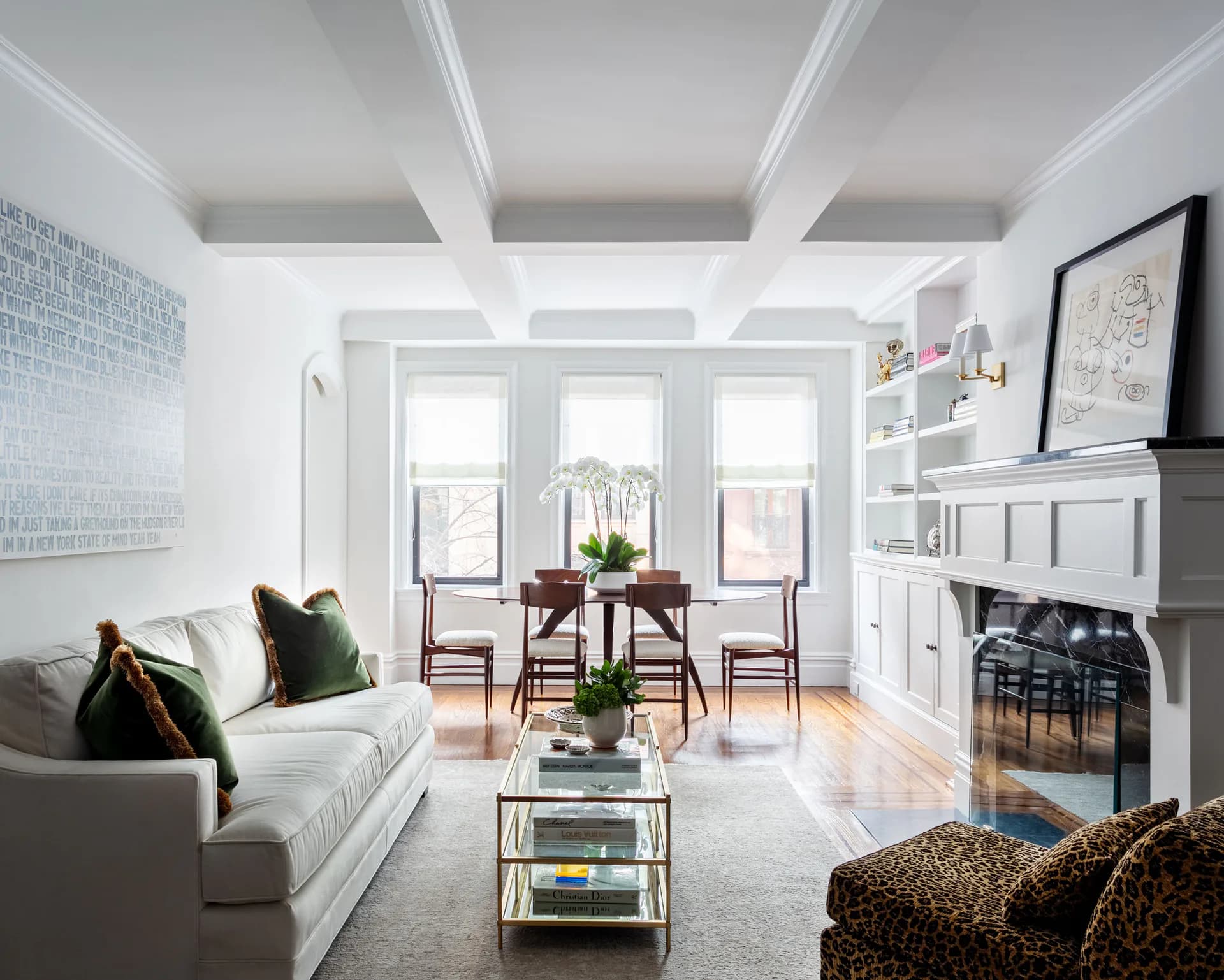 Bright, airy living room with coffered ceiling, fireplace, neutral seating, and large windows overlooking cityscape by Karen Loc.