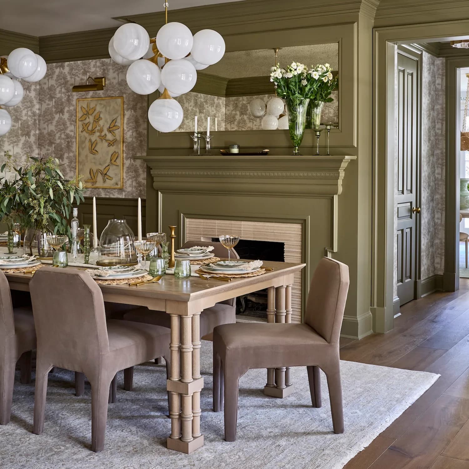 Elegant dining room with sage green shiplap walls, natural wood table, taupe upholstered chairs, fireplace, and sculptural white pendant lighting by Karen Wolf.