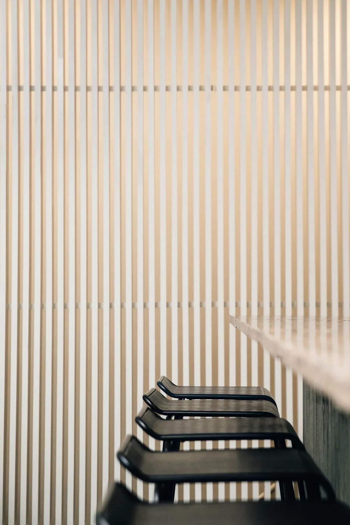 Minimalist interior with black lounge chairs positioned against a wall of vertical wooden slats creating rhythmic shadow patterns, by Kari Minami.
