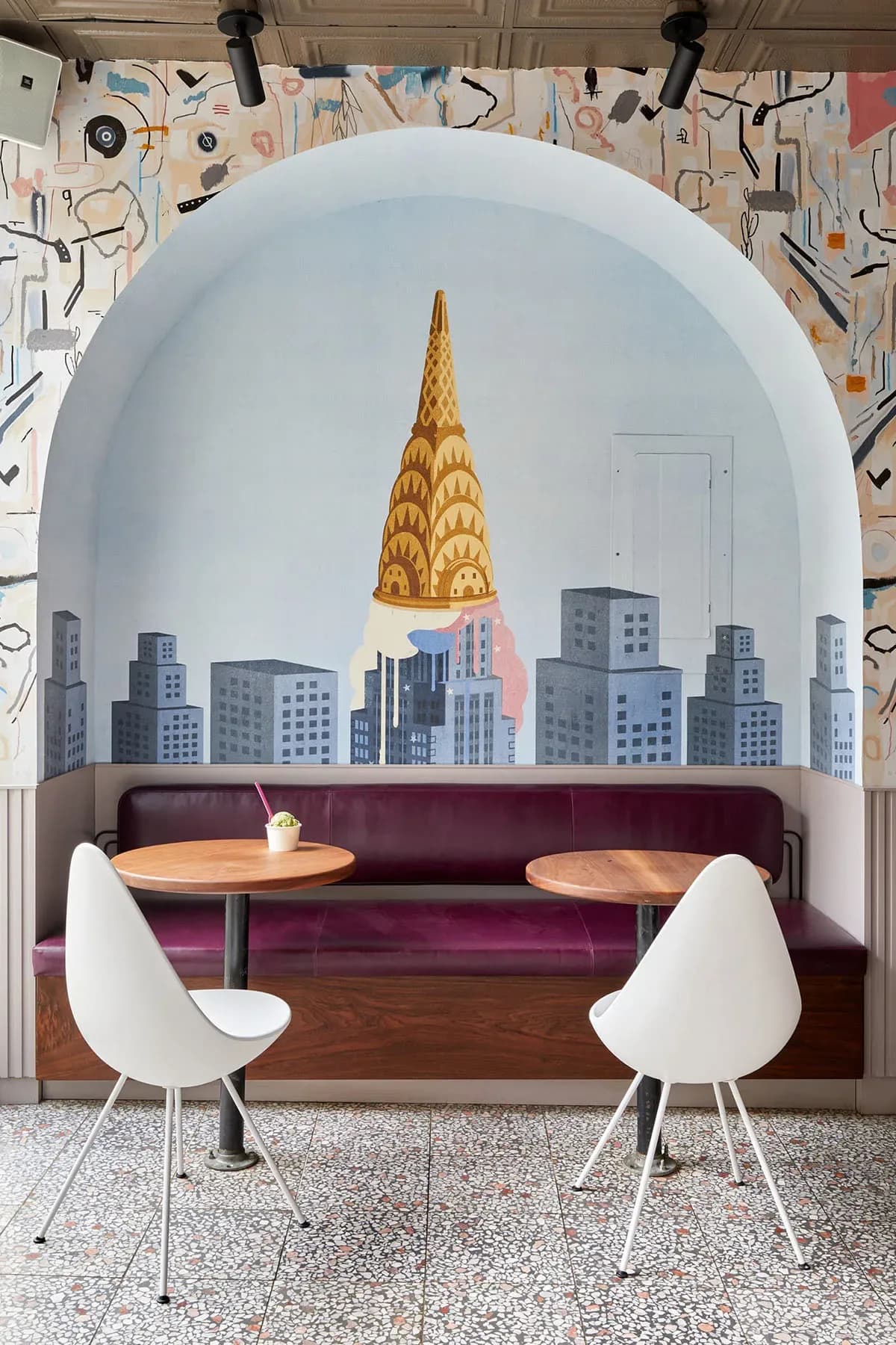 Modern booth seating with burgundy upholstery and round wood tables beneath a curved alcove featuring a Chrysler Building mural, by Kari Minami.
