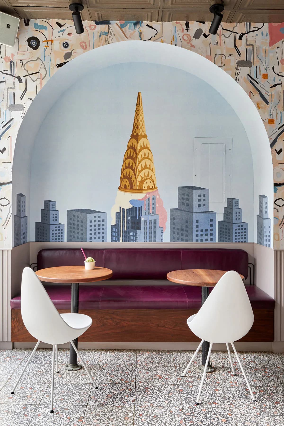 A modern booth seating area with burgundy upholstery and round wooden tables, white chairs, and a curved white alcove featuring a Chrysler Building mural and NYC skyline illustration by Kari Minami.