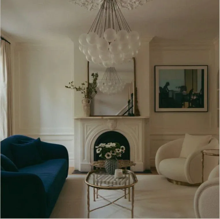 Contemporary living room with a navy curved sofa, cream armchair, round marble-top table, glass chandelier, fireplace with white surround, and neutral palette by Karla Goran.