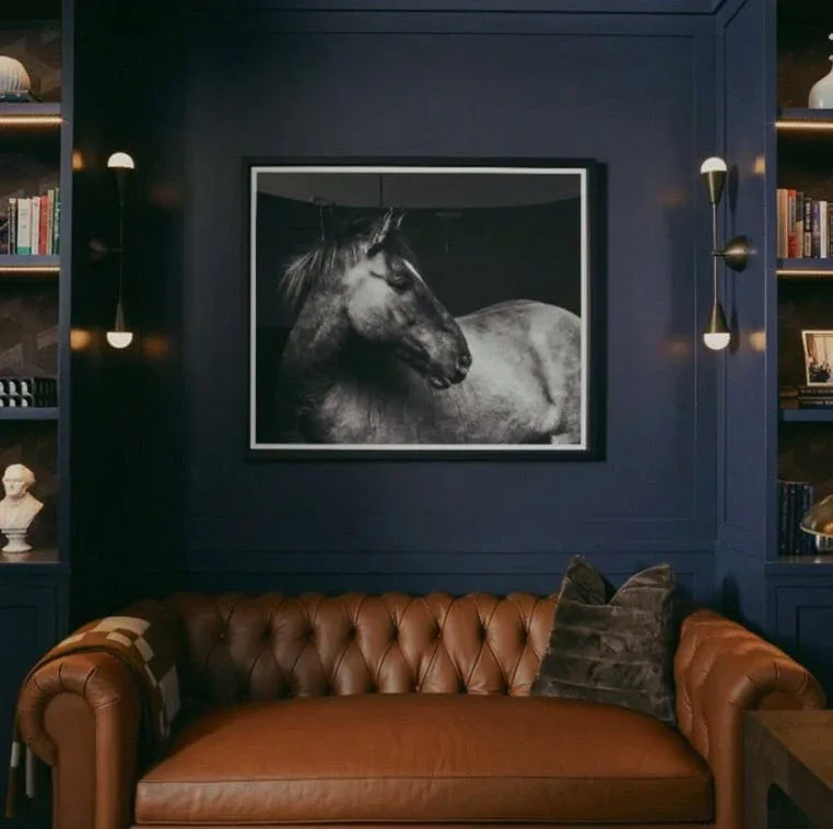 A luxurious navy blue interior with a cognac leather Chesterfield sofa beneath a black and white horse portrait, flanked by built-in shelving with brass sconces, by Karla Goran.