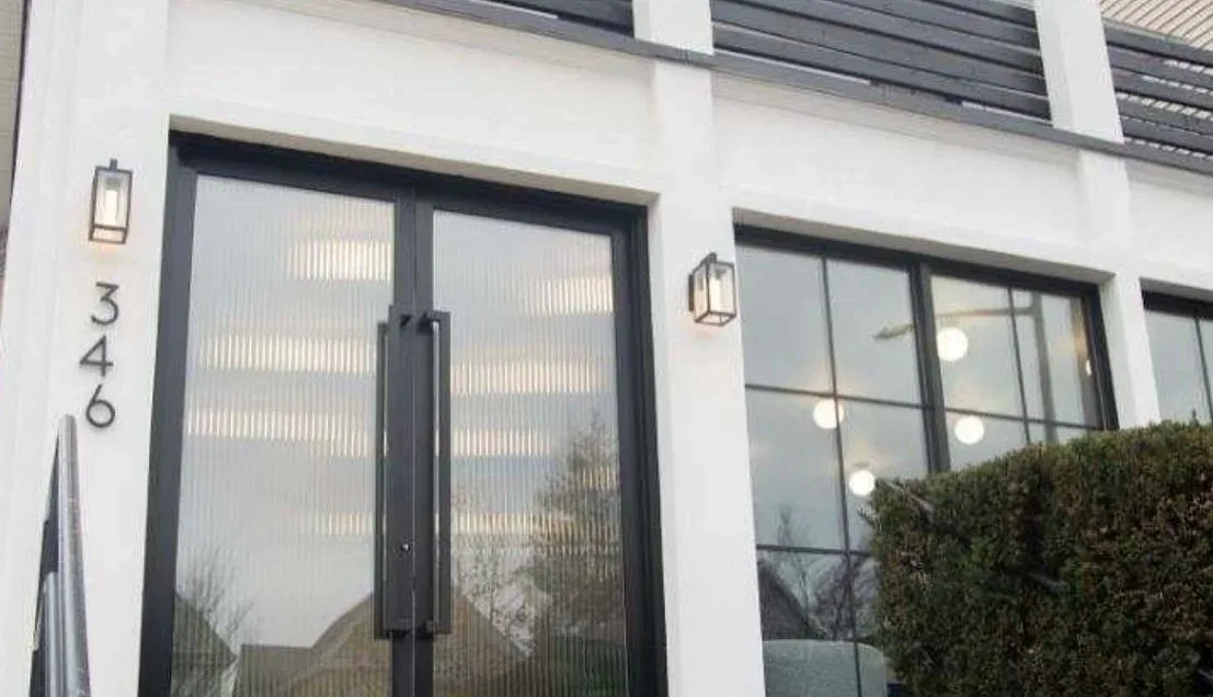 Modern white residential facade with black-framed glass doors and windows, brass sconces, and manicured hedging by Karla Quiroz.