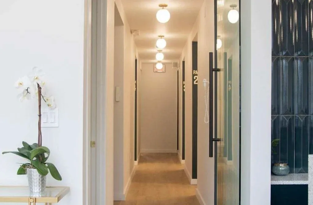 Modern hallway with warm wood flooring, white walls, recessed ceiling lights, and dark-framed glass doors lining both sides, designed by Karla Quiroz.