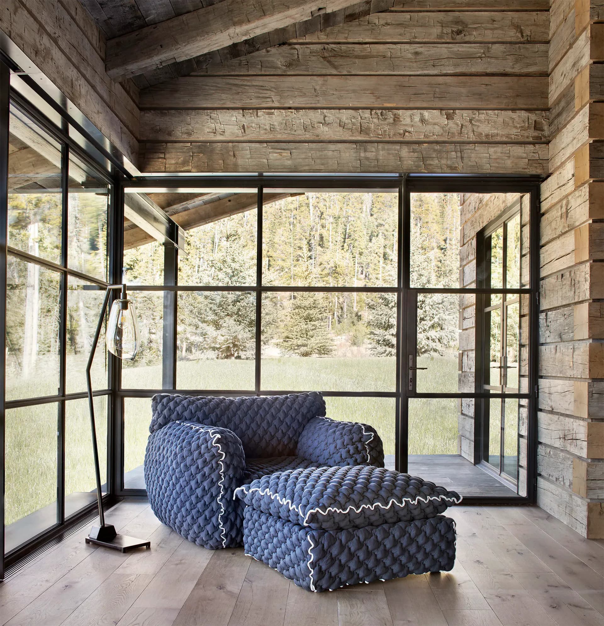Modern screened porch with exposed wooden beams, large gridded windows, blue upholstered armchair and ottoman on light wood flooring, overlooking forest by Kary Bell.