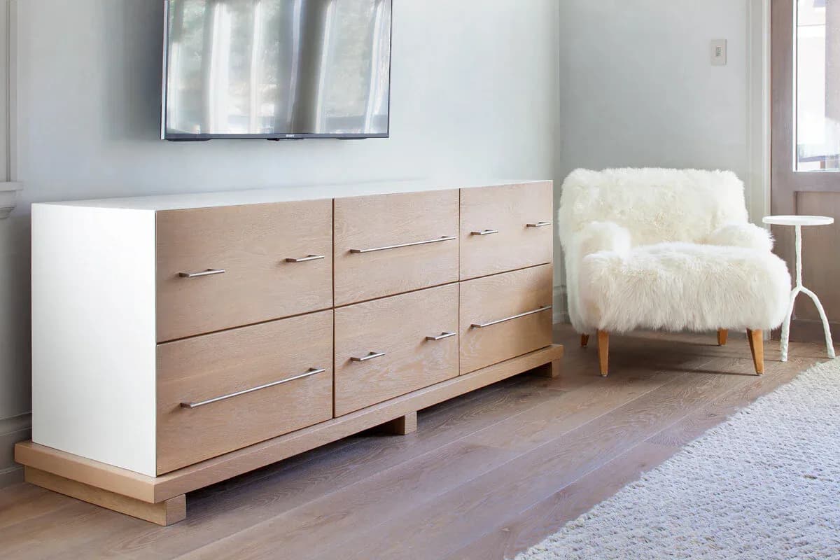 Modern bedroom interior featuring a light wood and white dresser with bronze handles, a cream-colored sculptural armchair, and neutral palette by Kary Bell.