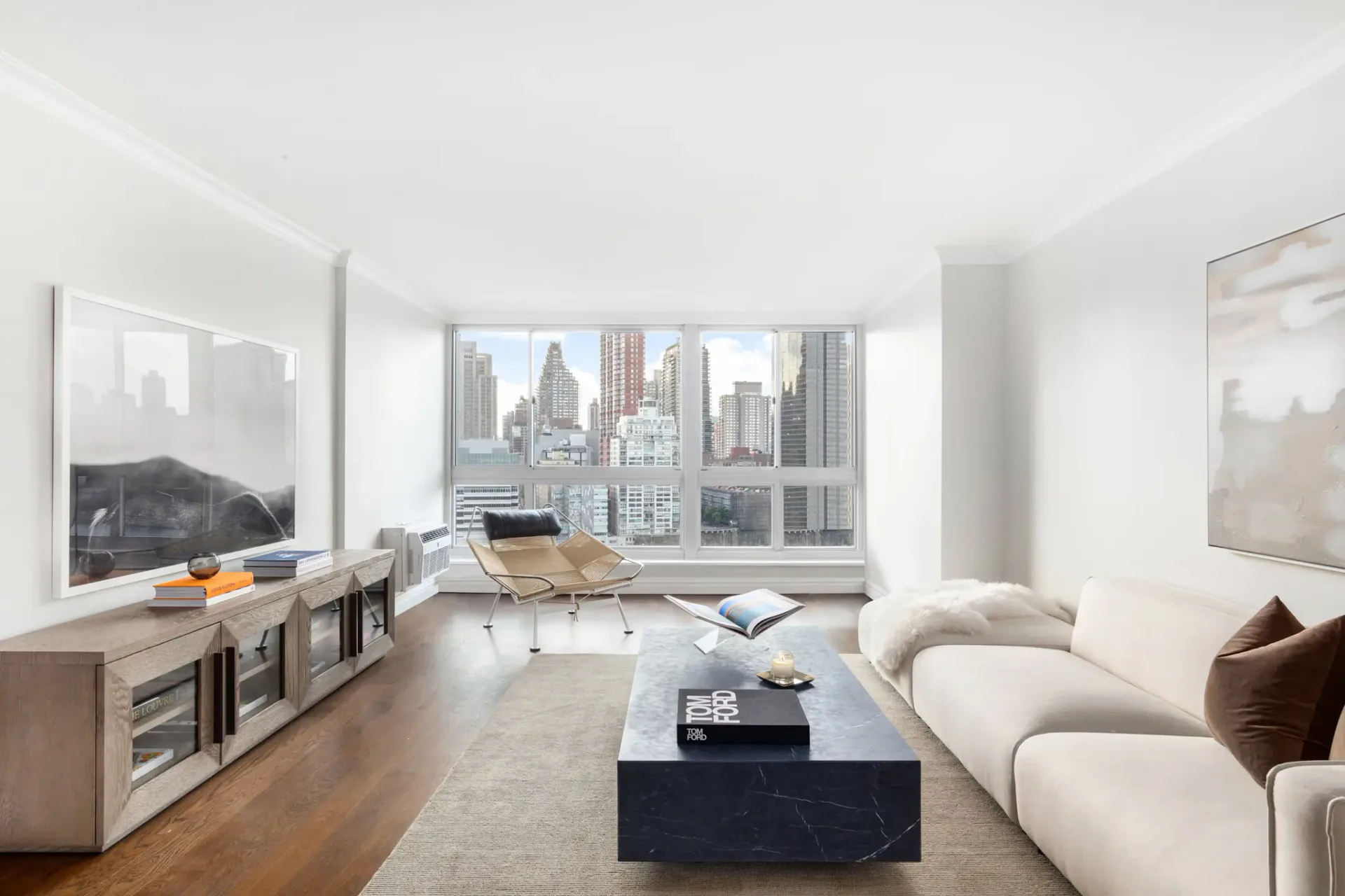 Modern high-rise living room with curved cream sofa, dark coffee table, wood media console, and floor-to-ceiling windows overlooking Manhattan skyline by Kate Cuadro.