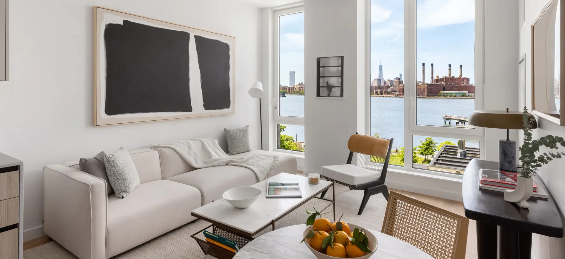 Bright modern living room with cream sofa, floor-to-ceiling windows overlooking water, abstract wall art, and mid-century furniture by Kate Cuadro.
