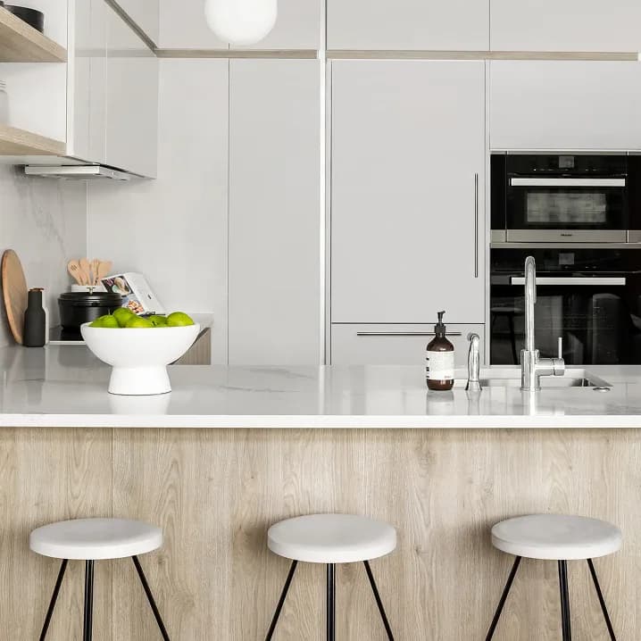 Modern white kitchen with light wood base, white quartz countertop, built-in black appliances, and three white bar stools by Kate Cuadro.