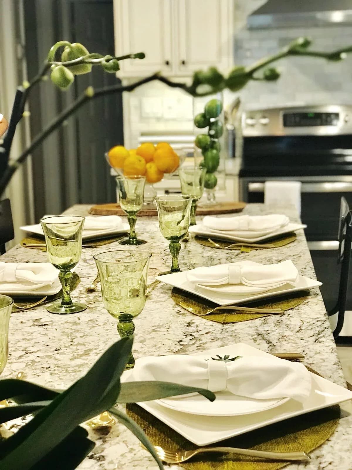 Elegant dining table with marble countertop, green glassware, white place settings, and yellow floral centerpiece in a modern kitchen by Katherine Flores.