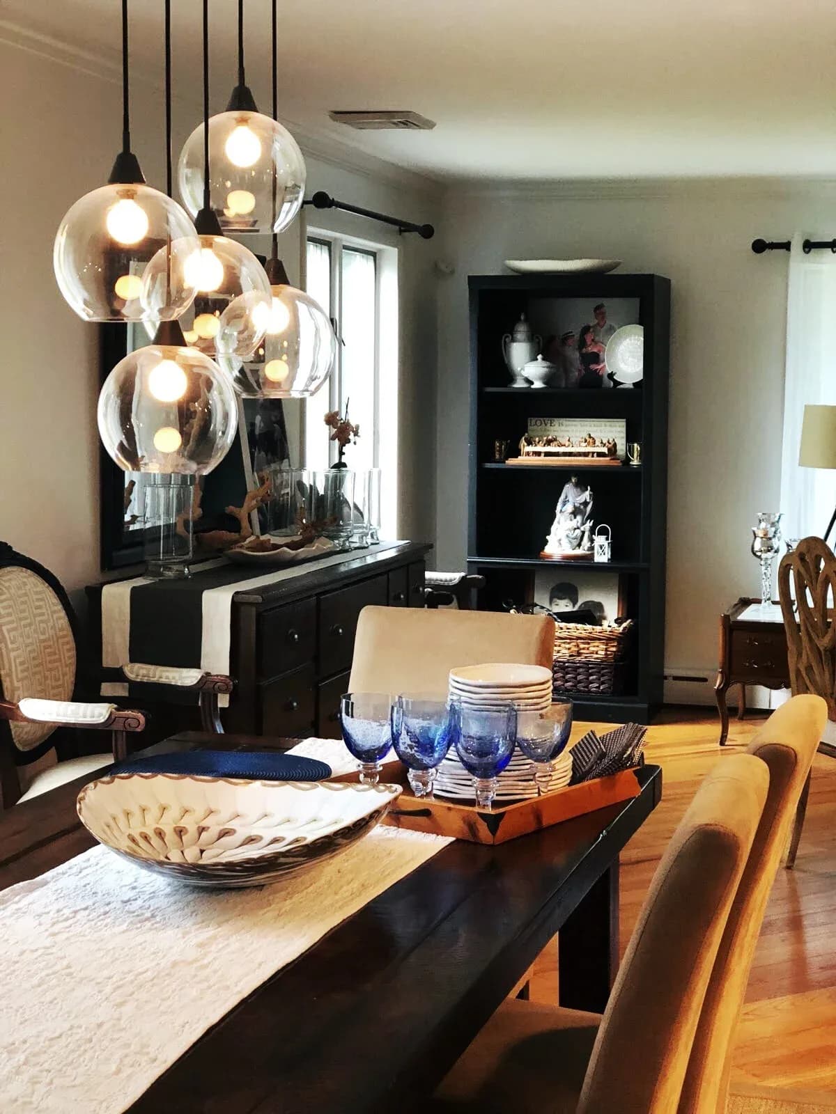 Modern dining room with black table, warm wood chairs, and sculptural glass pendant lights above; black shelving unit with decorative objects and fireplace insert by Katherine Flores.
