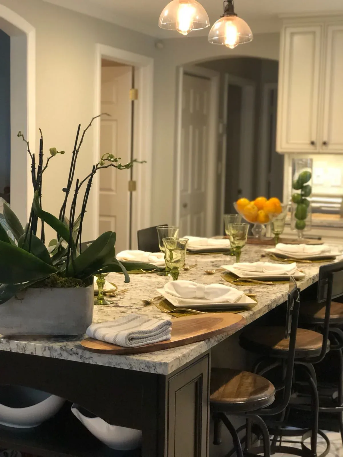 Modern kitchen dining area with marble countertop island, black cabinetry, white subway tile backsplash, pendant lighting, and place settings with yellow flowers by Katherine Flores.