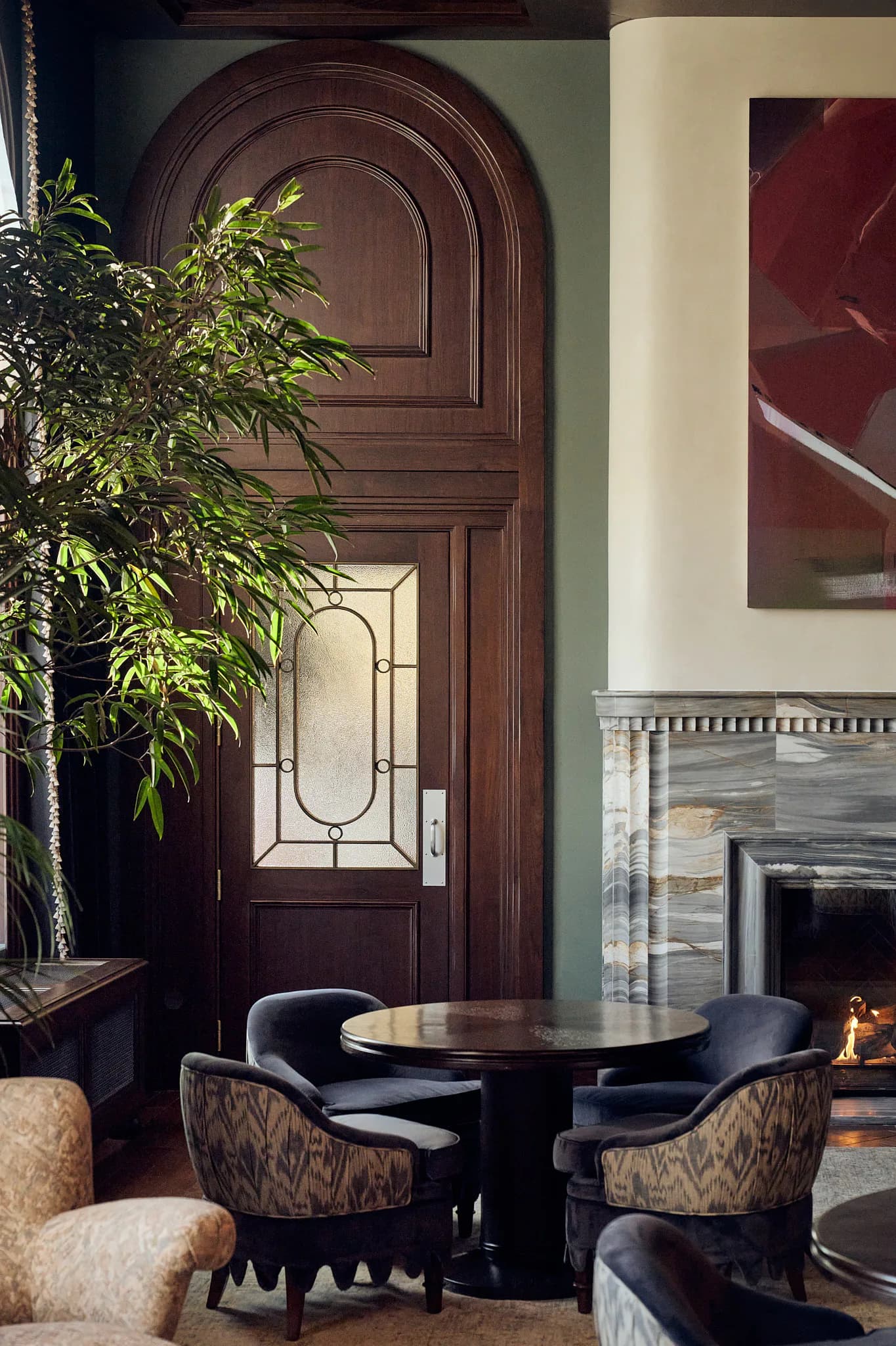 Modern living room with dark wood arched doorway, round black table with patterned chairs, green plant, fireplace, and sage green walls by Katie Cole.