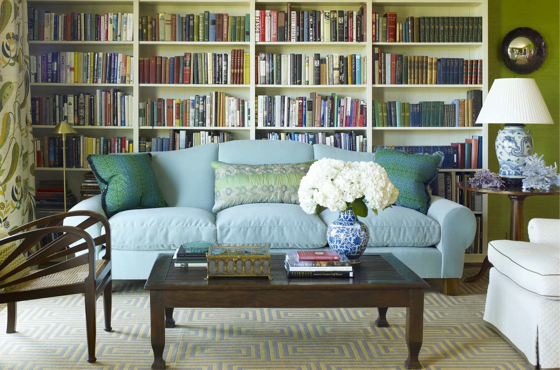 Light blue upholstered sofa with green and blue pillows positioned before floor-to-ceiling bookshelves, styled with dark wood coffee table and white flowers, designed by Katie Ridder.