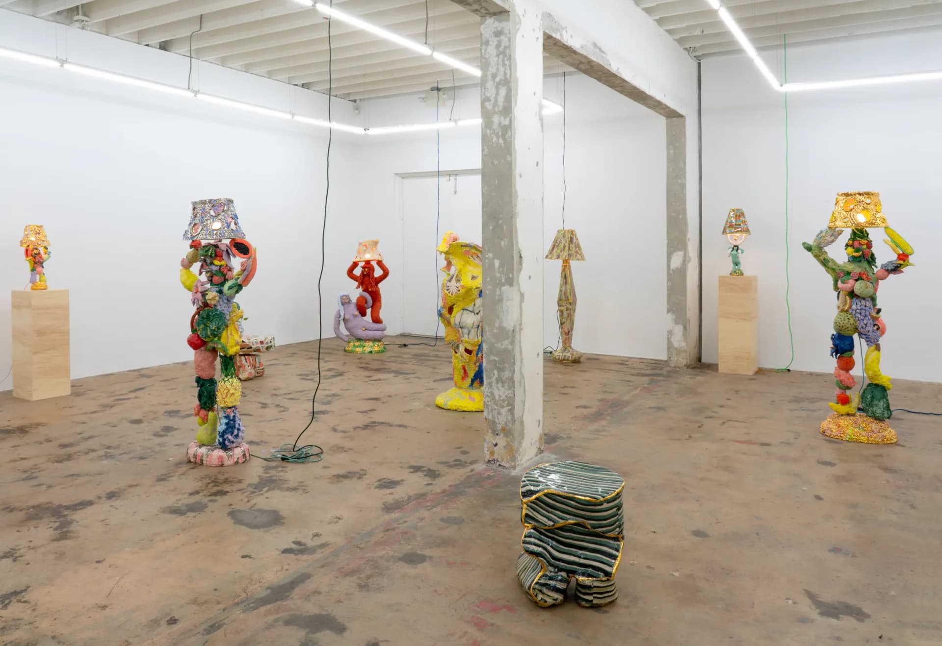 Industrial gallery space with suspended sculptural installations made from stacked colorful objects and wooden pedestals, concrete floor, by Katie Stout.