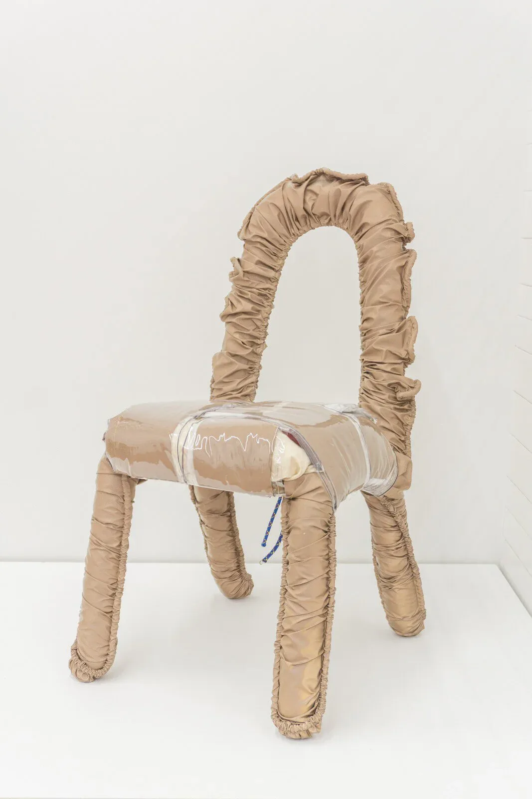 Beige upholstered chair with gathered fabric detailing and curved backrest, featuring clear plastic seat wrap, by Katie Stout.