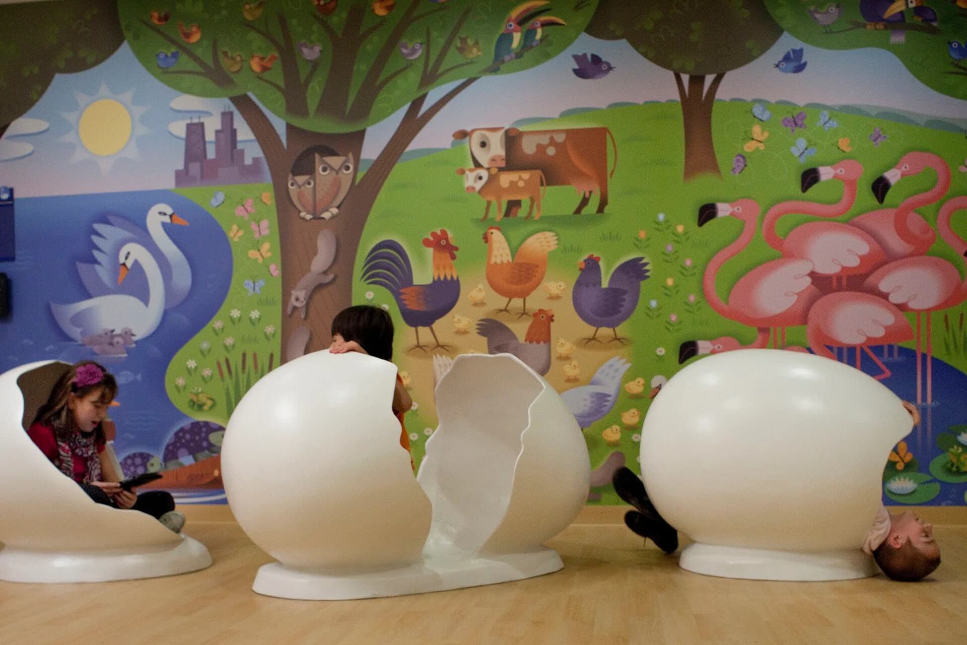 Three white sculptural egg-shaped installations displayed in front of a colorful children's mural featuring farm animals and nature scenes by Kaylie Siwy.