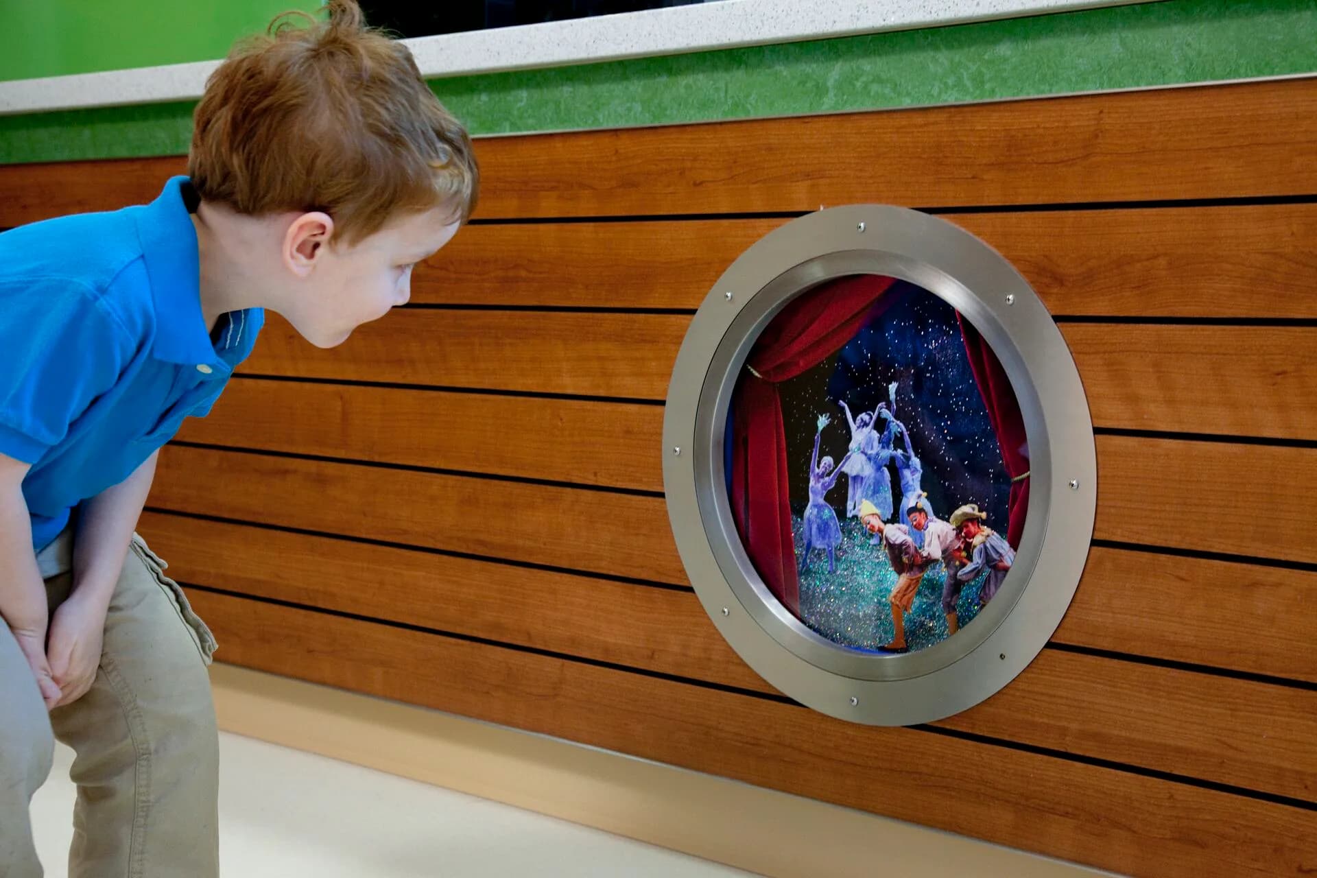 Child viewing a circular porthole window installation with theatrical curtains and figures inside, mounted on wooden wall paneling by Kaylie Siwy.