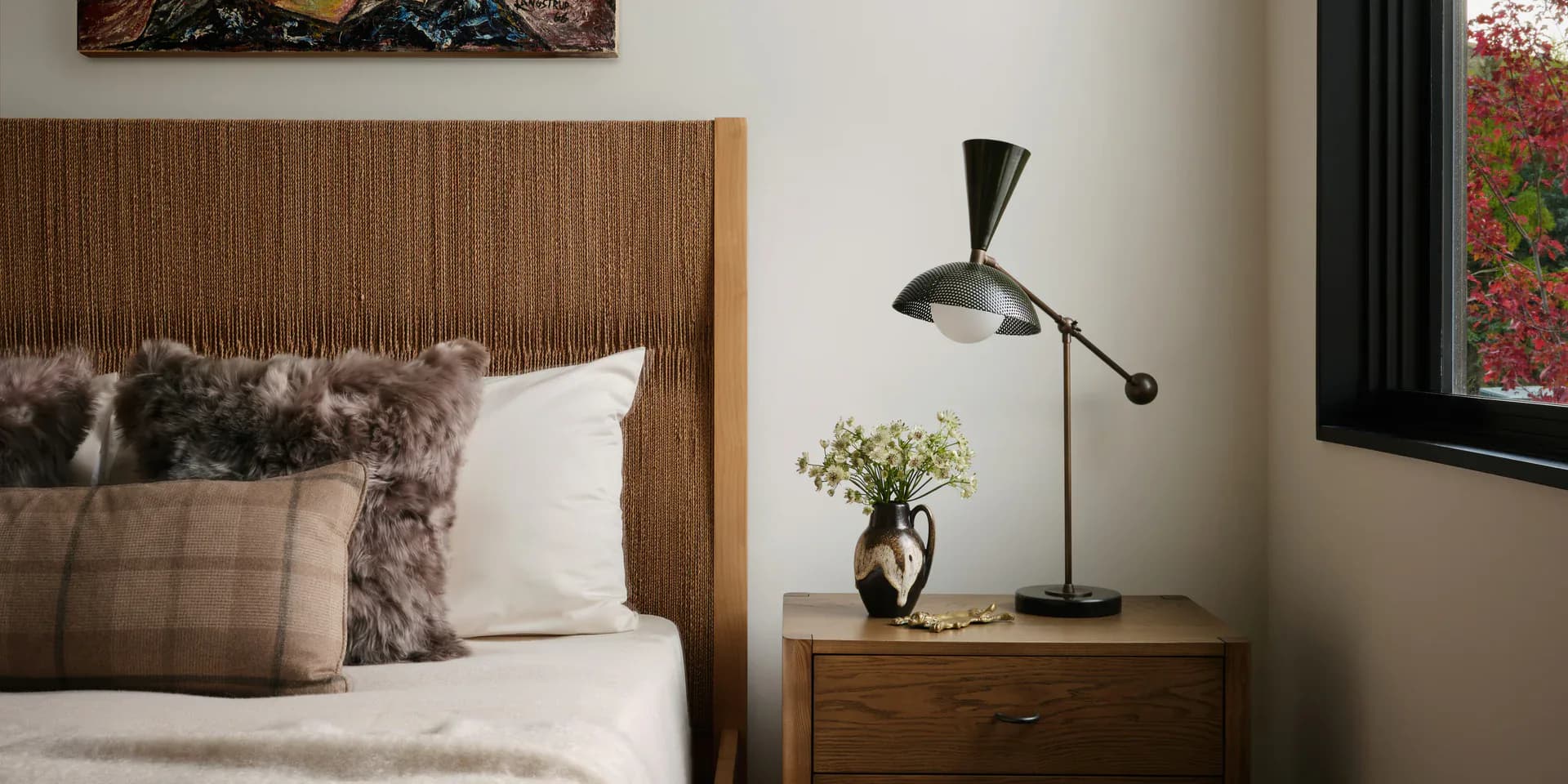Modern bedroom with ribbed wooden headboard, layered neutral textiles, wooden nightstand with black task lamp and potted flowers, and framed artwork by Kelly Aaron.
