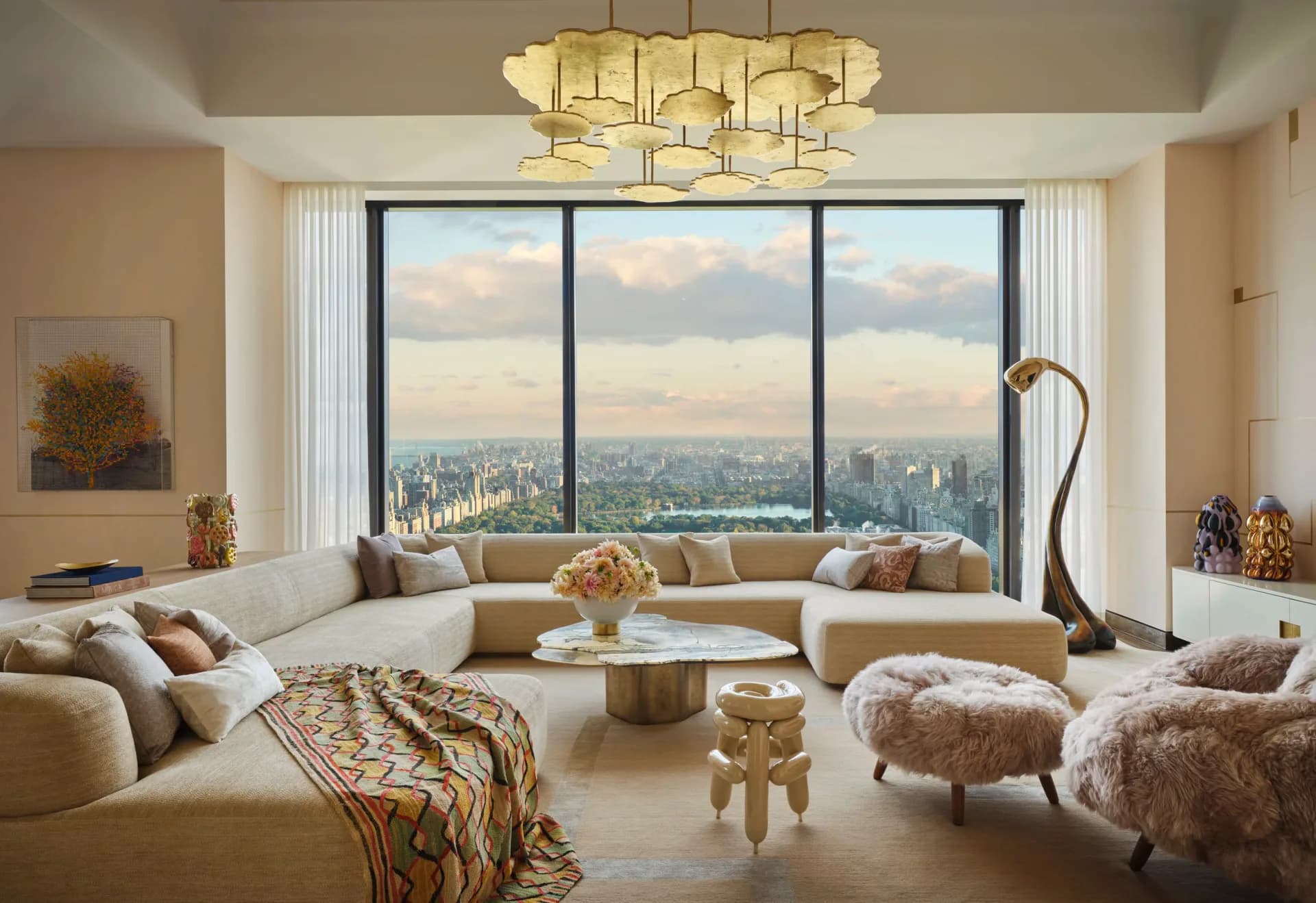 Luxurious high-rise living room with curved cream seating, panoramic city skyline views, brass pendant lighting, and warm neutral palette by Kelly Behun.