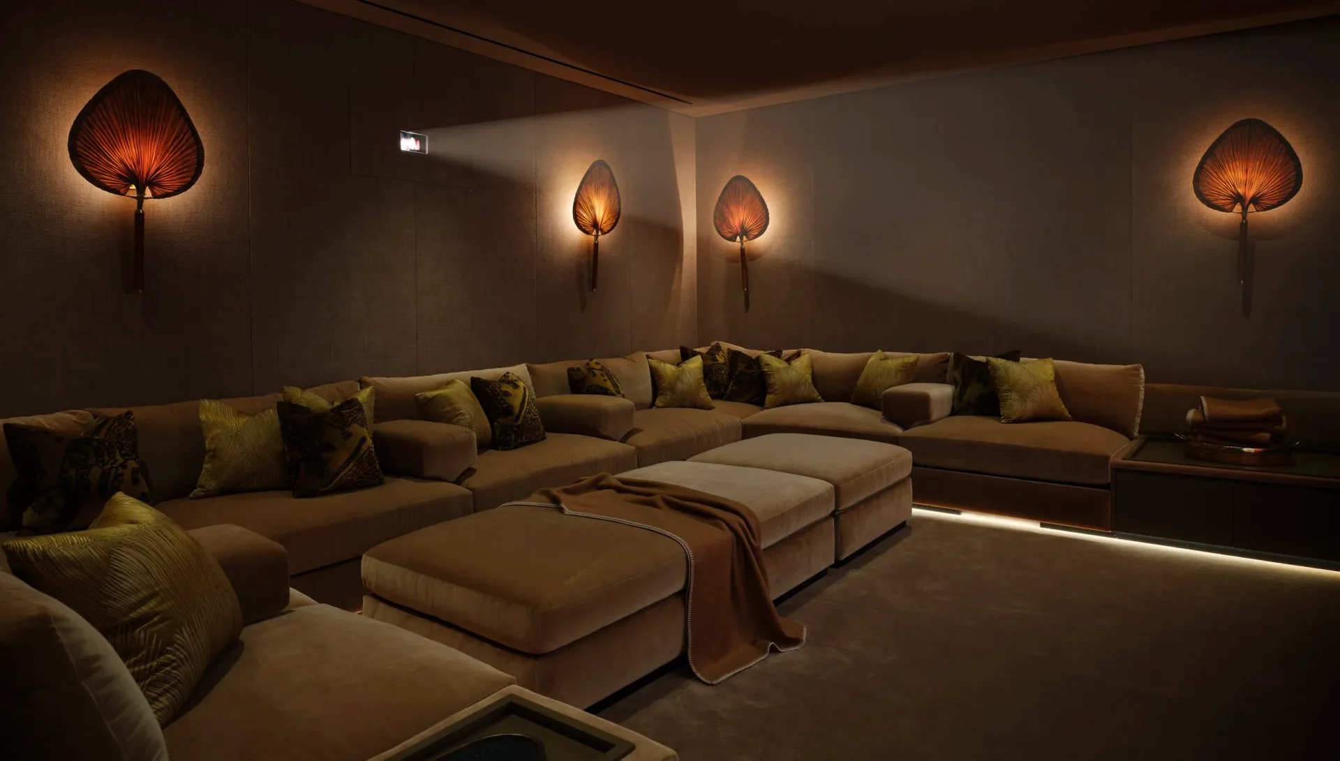 Luxurious media lounge with curved sectional seating, warm ambient lighting from decorative wall sconces, and glowing floor accent lighting by Kelly Behun.