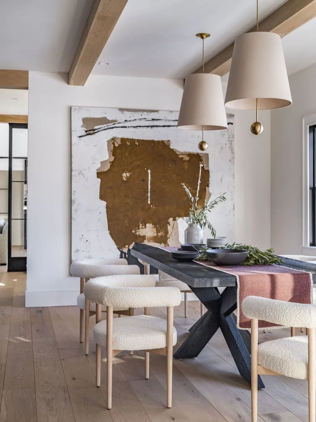 Modern dining room with black X-base table, cream upholstered chairs, abstract gold artwork, wooden beams, and pendant lights by Kelly Mittleman.