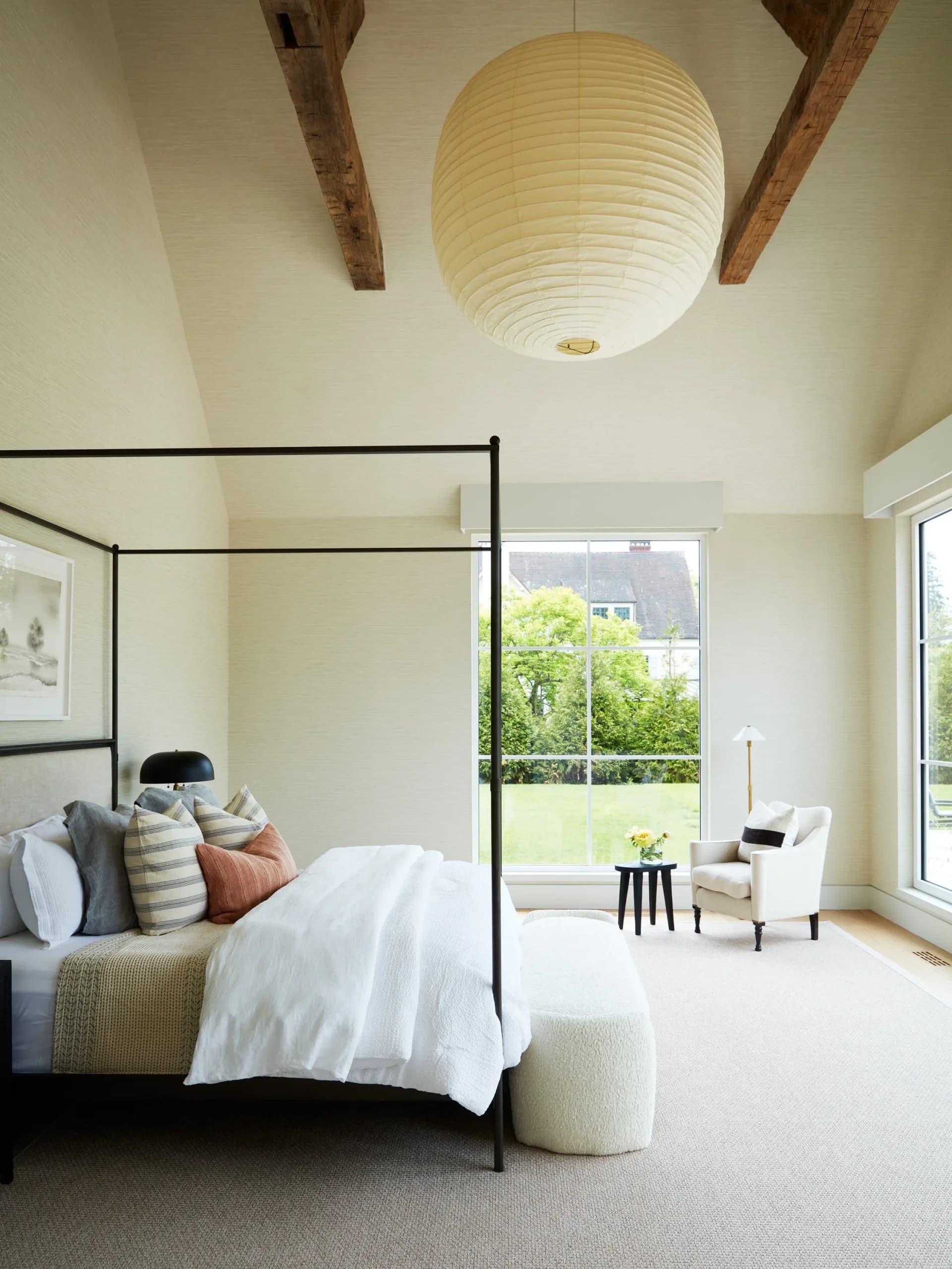 Serene primary bedroom features a black four-poster bed, woven pendant light, wooden ceiling beams, and expansive windows overlooking greenery by Kelly Mittleman.