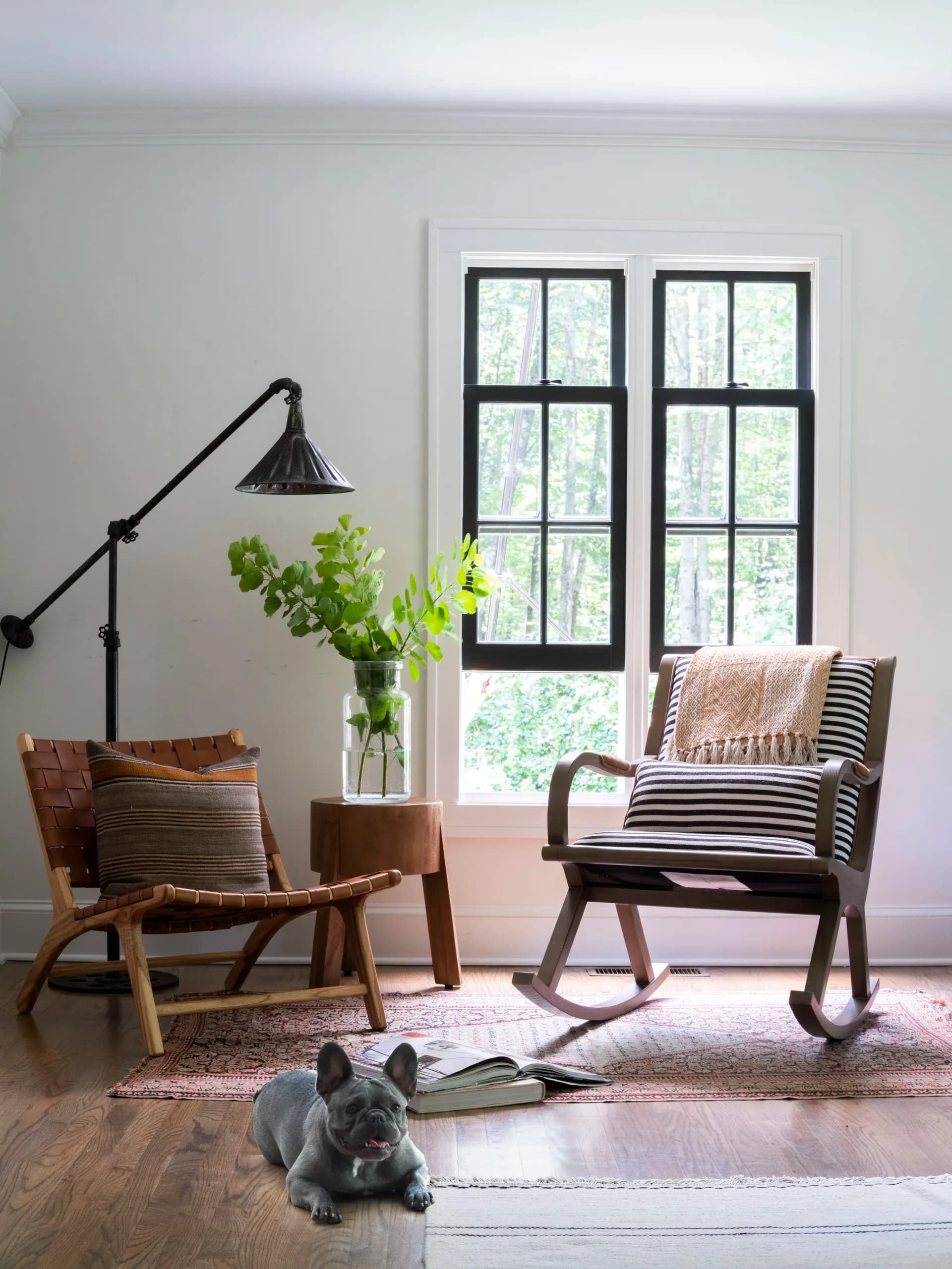 Minimalist living room with mid-century modern wooden chairs, black arc lamp, tall windows with black frames, fresh greenery, and striped textiles by Kelly Mittleman.