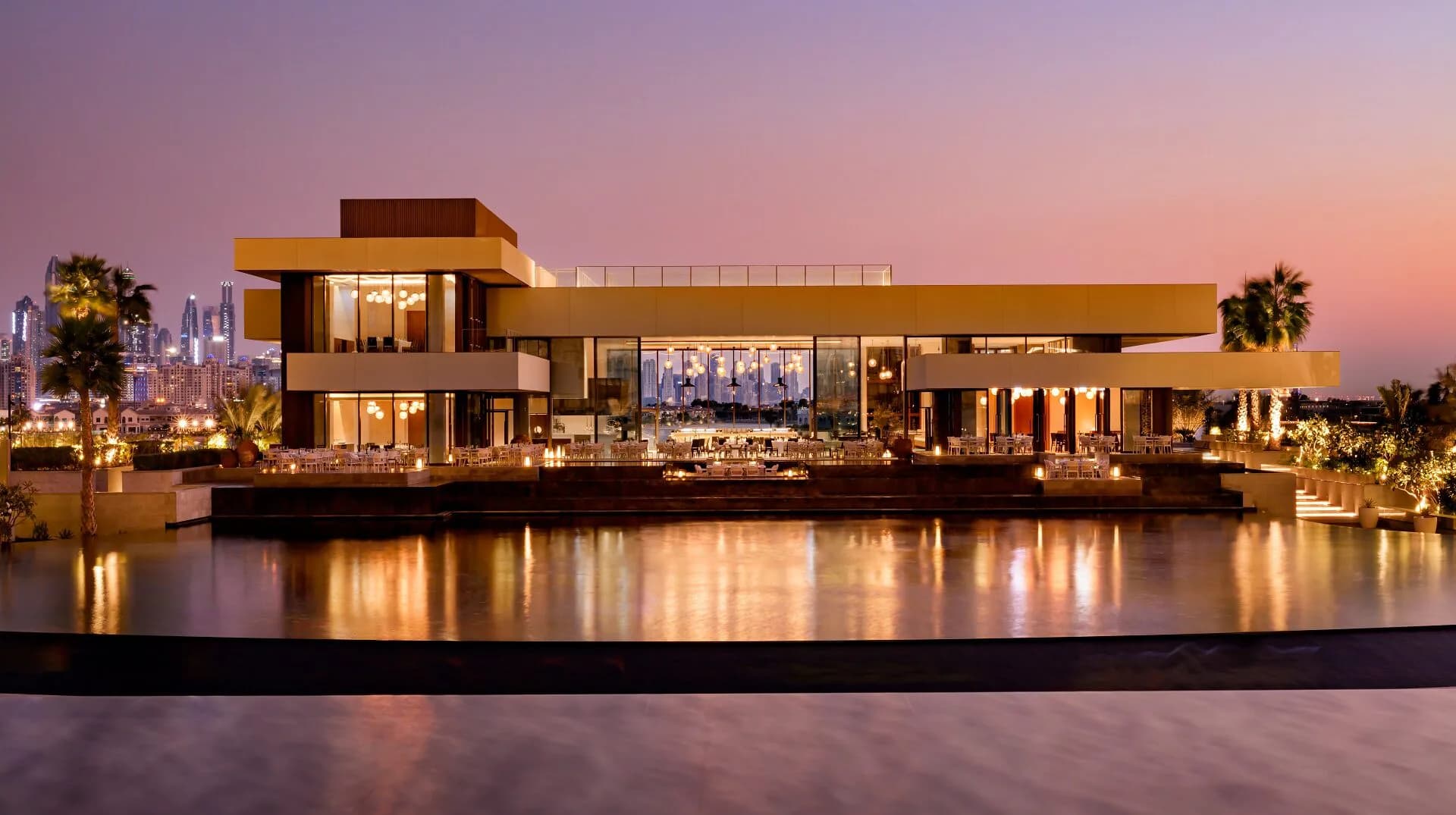 Modern waterfront residence with warm golden lighting at dusk, featuring glass walls, clean lines, and reflections in still water by Kelsey Verillo.
