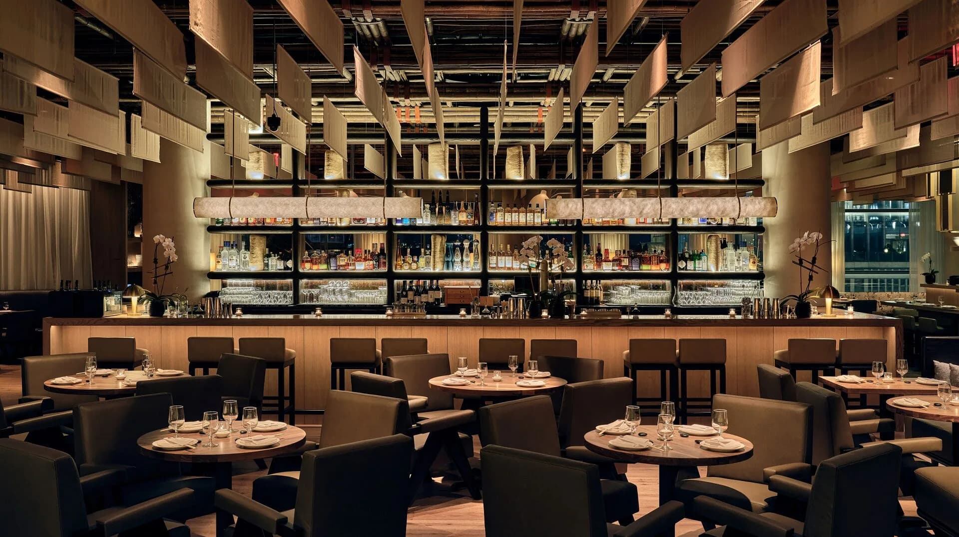 Upscale restaurant interior with warm wood tones, curved banquette seating, round tables, and illuminated shelving wall by Kelsey Verillo.