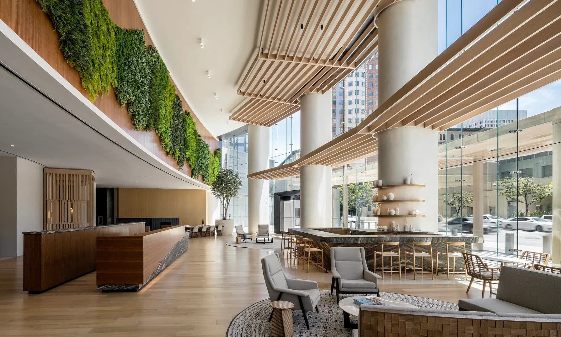 Modern hotel or corporate lobby with soaring ceilings, wooden slat details, living green walls, and floor-to-ceiling windows overlooking city views by Kelsey Verillo.
