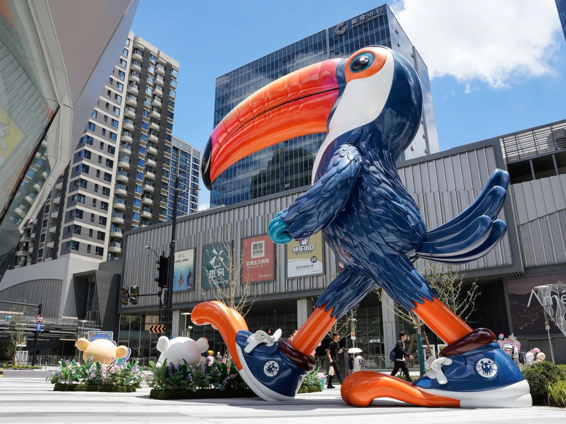 Large-scale public art installation featuring a stylized toucan sculpture in orange and navy blue, positioned in an urban plaza surrounded by modern buildings by Ken Kelleher.