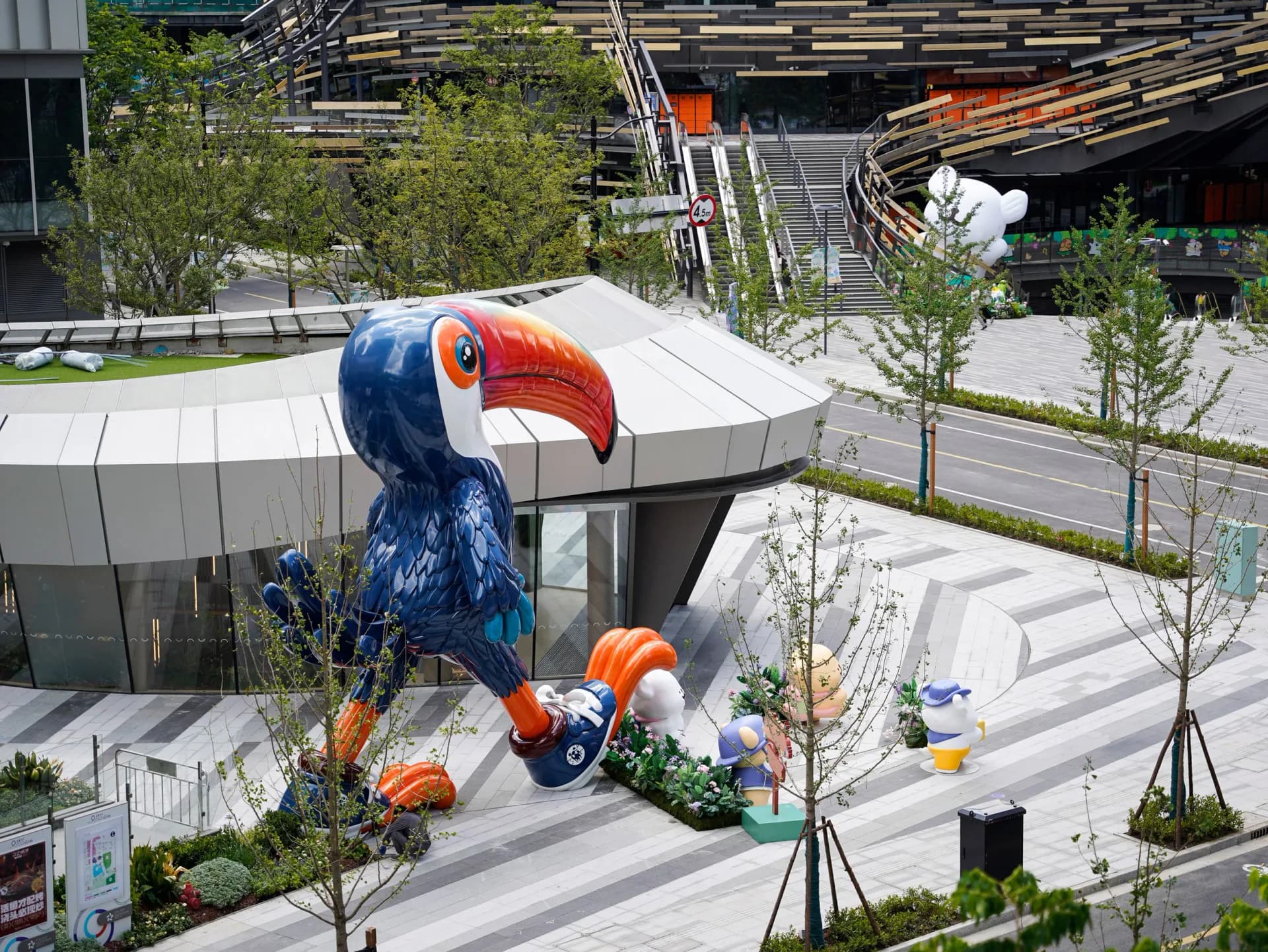 Aerial view of modern public plaza with sculptural blue and orange bird installation, white curved building, landscaping, and pedestrian pathways by Ken Kelleher.