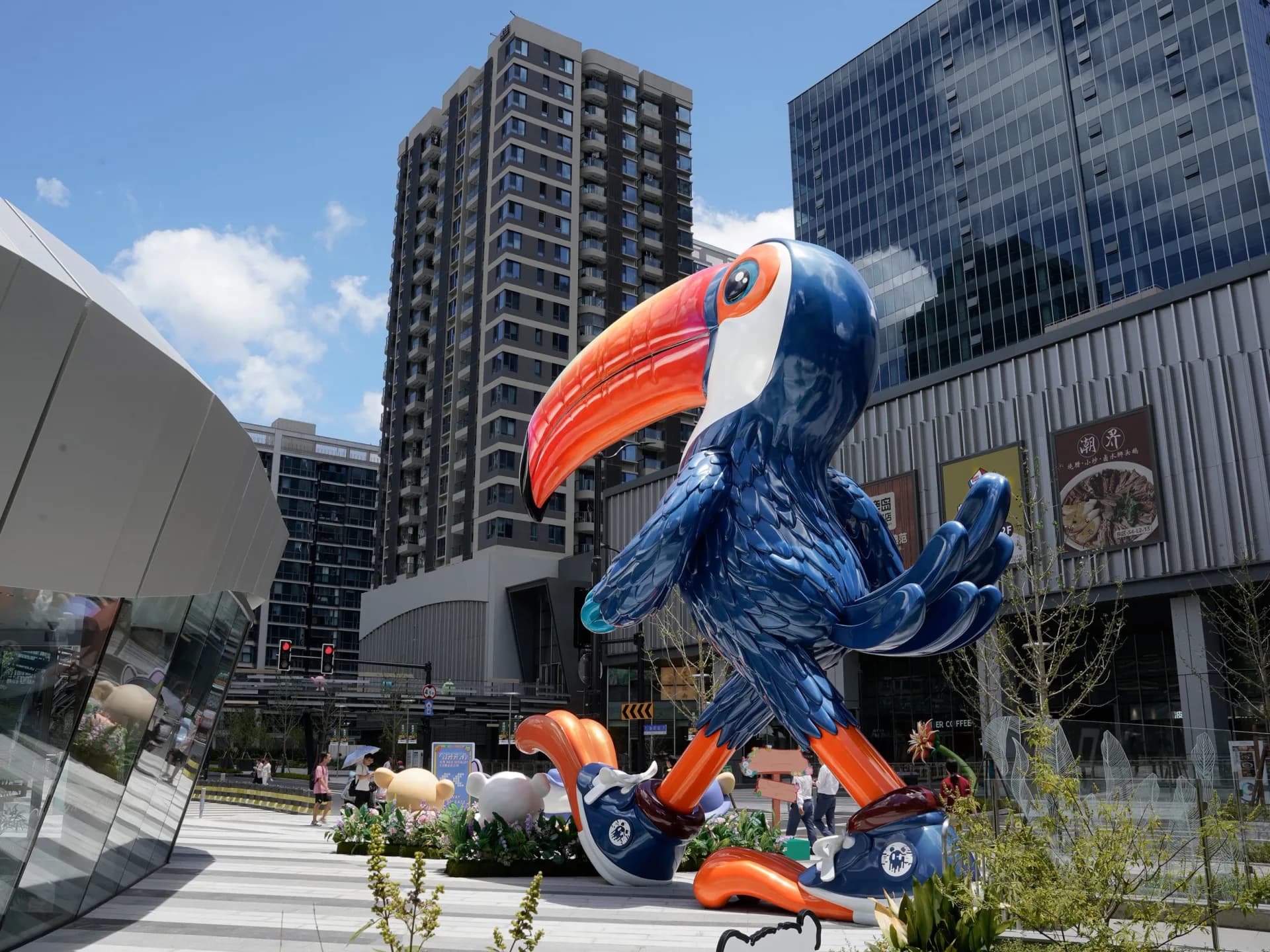 Large colorful blue and orange sculptural toucan installation in an urban plaza with contemporary buildings and landscaping by Ken Kelleher.