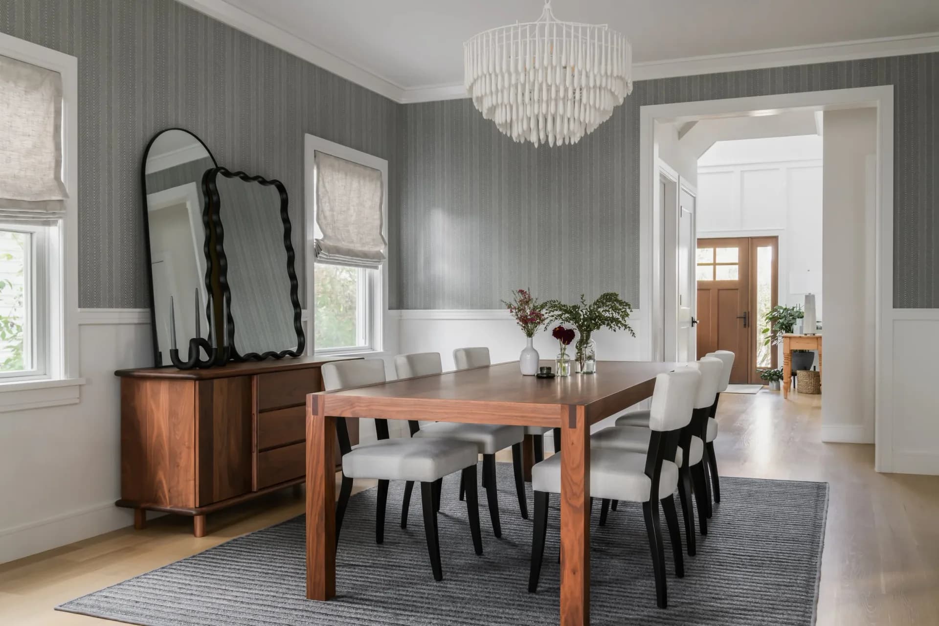 A modern dining room features a wooden table with black and white chairs, gray striped wallpaper, white wainscoting, and a crystal chandelier by Keri Tickle.