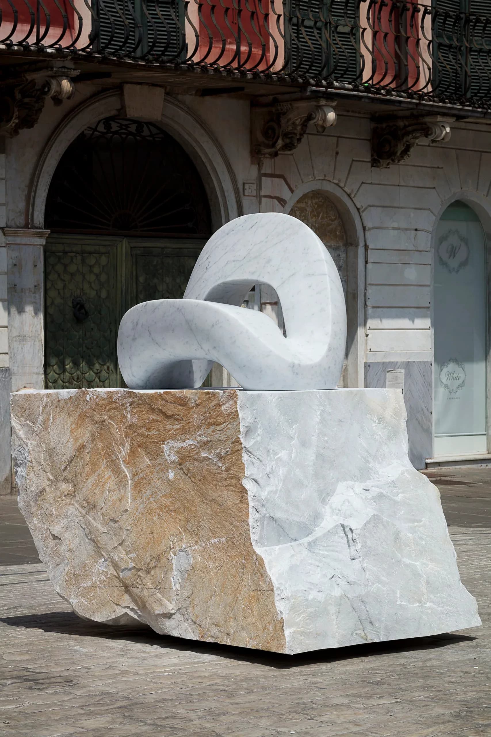 White marble sculptural installation with curved organic forms mounted on a tan stone base, displayed in front of a classical building facade by Kickie Chudikova.