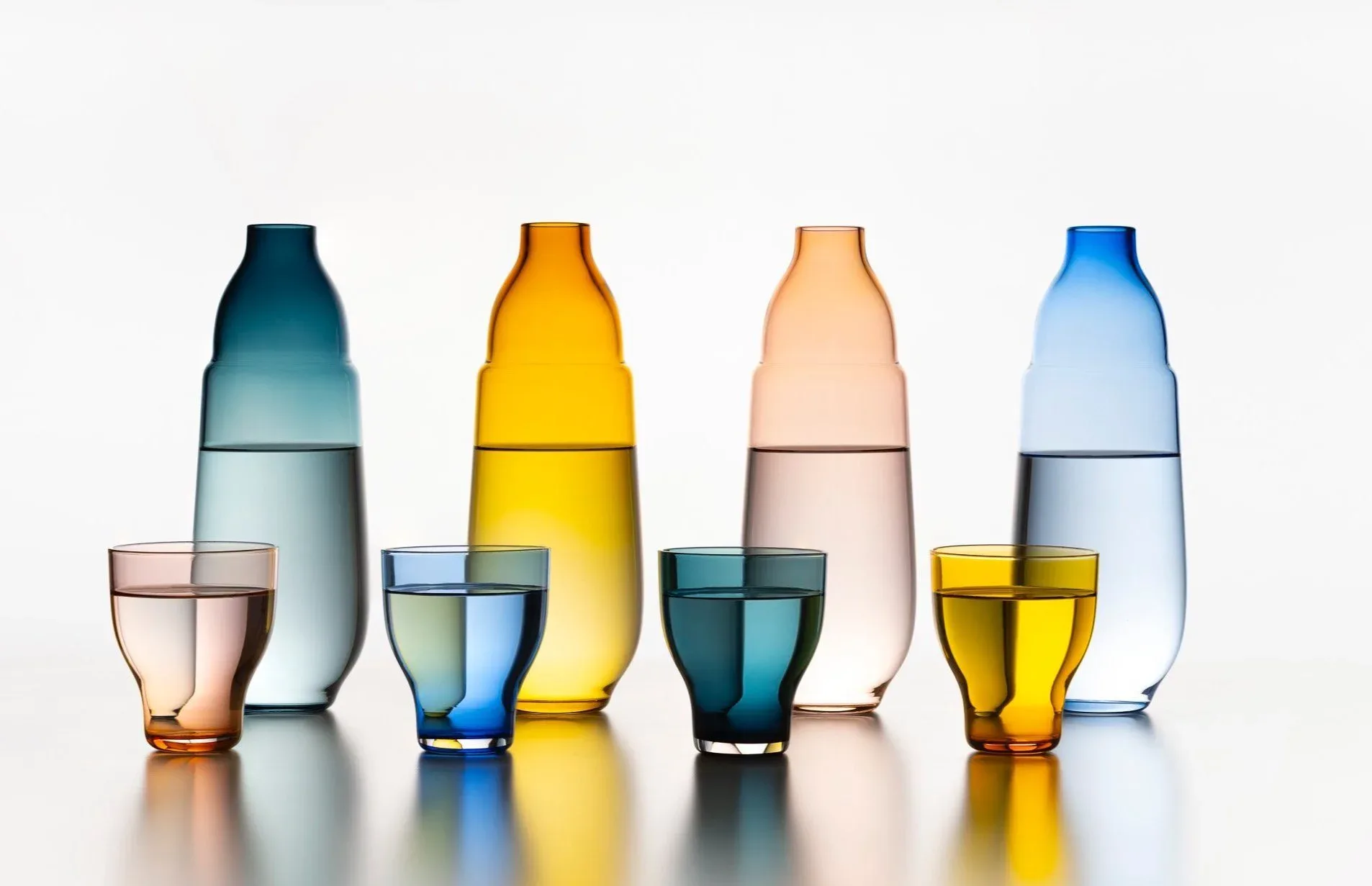 Four colorful glass bottles with matching drinking glasses containing tinted water in teal, yellow, peach, and blue hues by Kickie Chudikova.