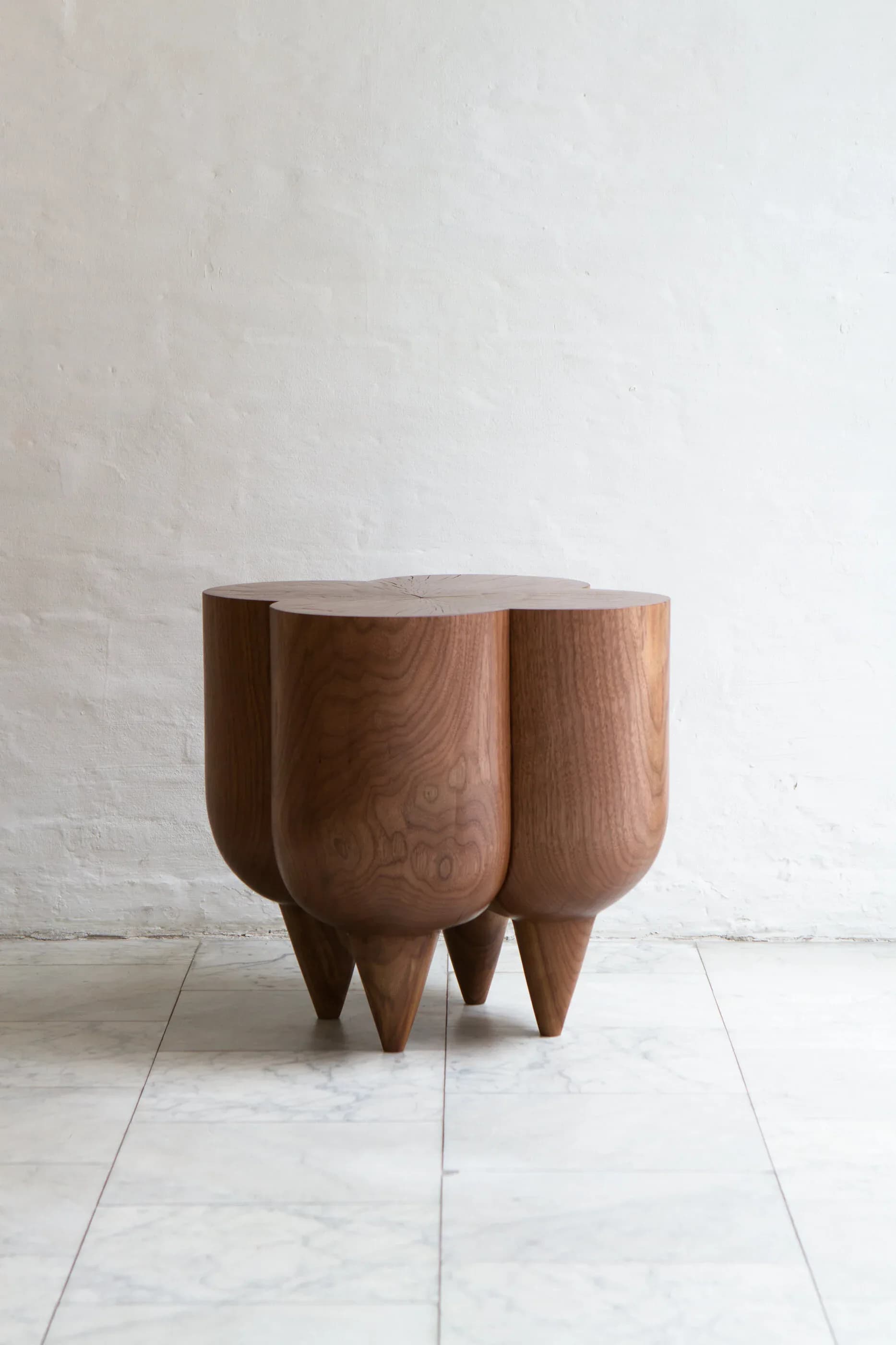 A sculptural wooden side table with rounded, organic forms and tapered legs in warm walnut, photographed against white walls and marble flooring by Kieran Kinsella.