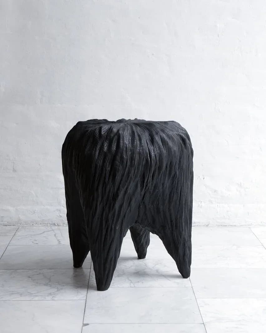 Black sculptural stool with dramatic pleated vertical ridges and splayed legs on white tile floor against white brick wall by Kieran Kinsella.