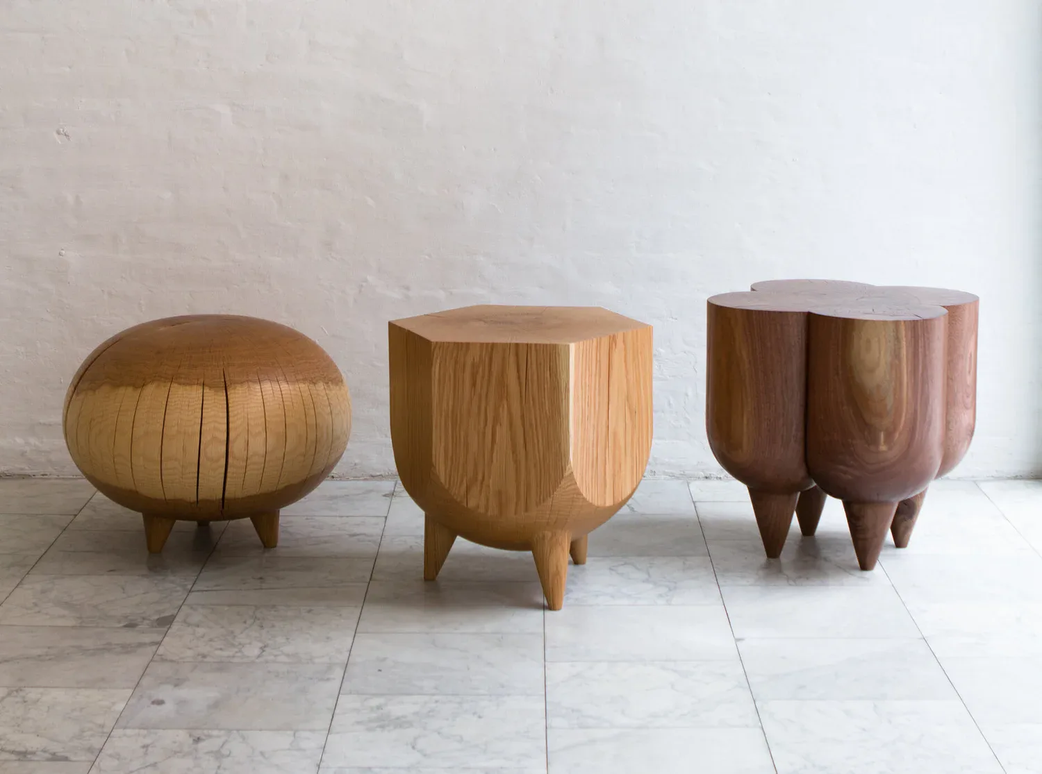 Three sculptural wooden stools with organic forms displayed on pale tile flooring against a white wall, showcasing furniture design by Kieran Kinsella.