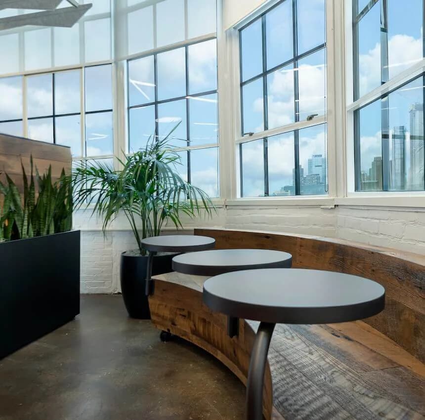 Modern office lounge with round white-topped tables on wooden bases, black planters with greenery, and expansive grid windows overlooking a city skyline by EunHea Kim.